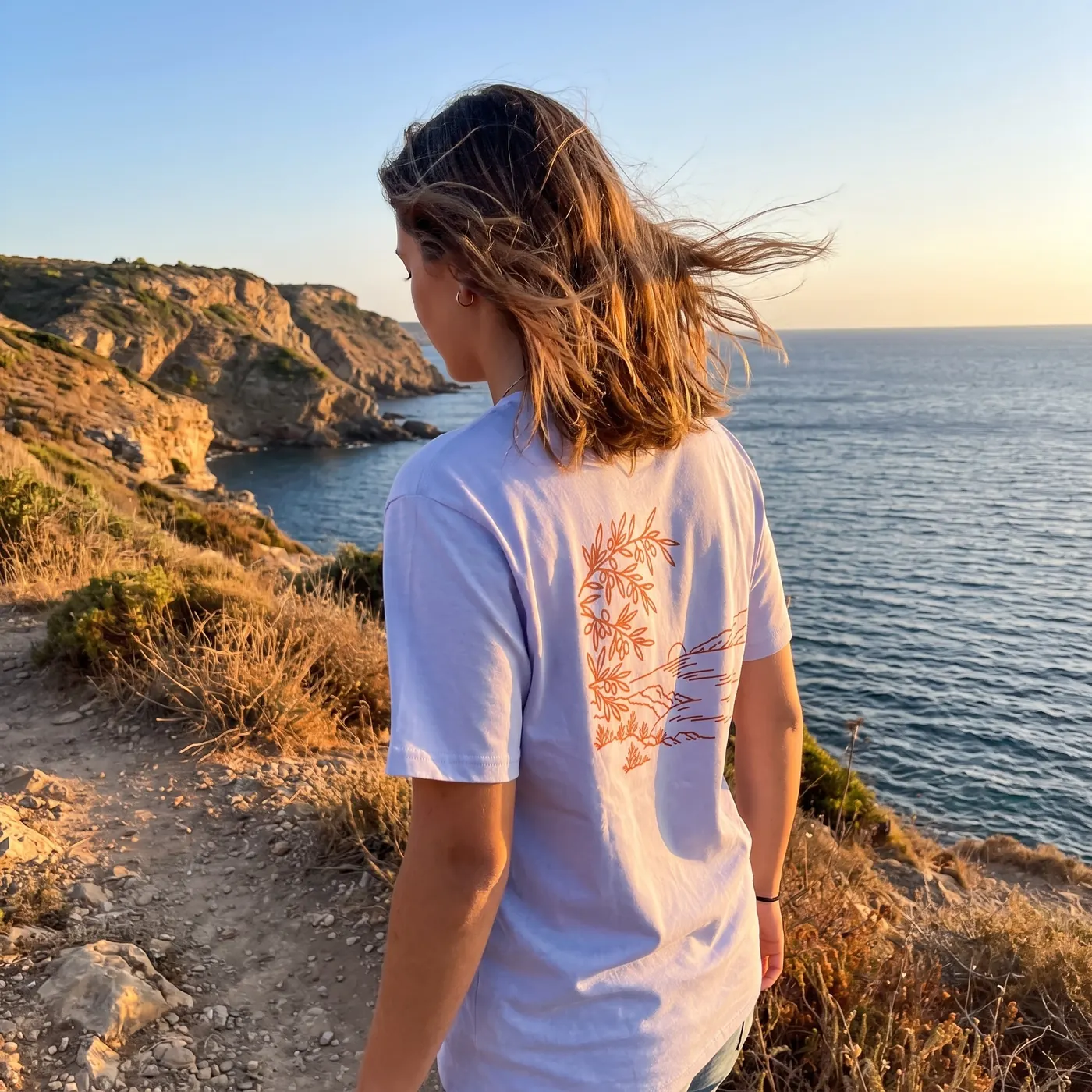 Au bord d'une calanque au soleil couchant, le dos du t-shirt Coucher de Sud montre son illustration imprimée